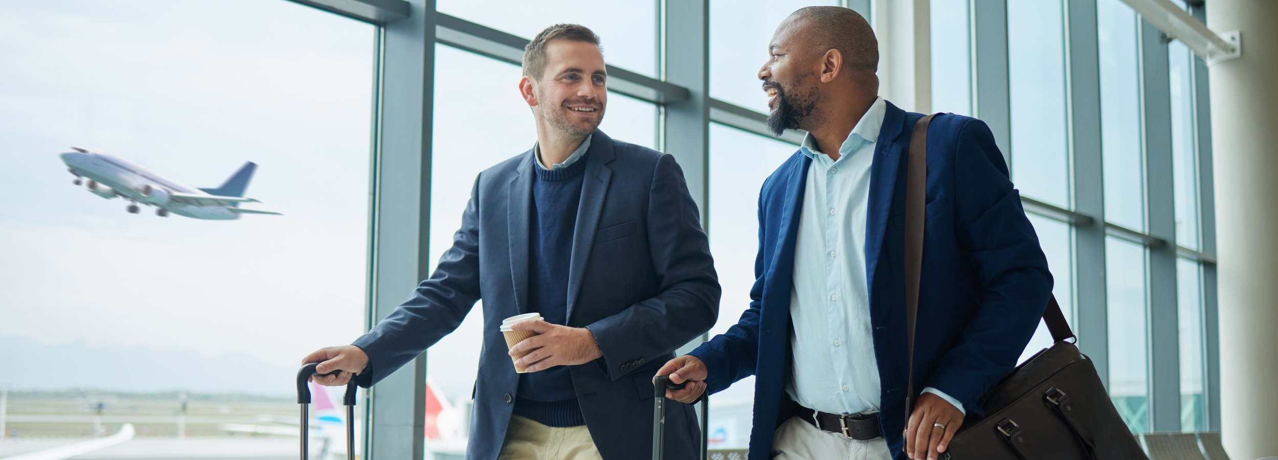Zwei Männer in Anzügen unterhalten sich lächelnd in einem Flughafen, während im Hintergrund ein Flugzeug startet.

Das Bild zeigt zwei Männer, vermutlich Geschäftspartner, die sich in einem modernen Flughafen befinden. Der Mann links hat hellbraunes Haar und ist in einen dunkelblauen Pullover unter einem marineblauen Blazer gekleidet, dazu trägt er helle Hosen. Er hält einen Pappbecher in seiner Hand und zieht einen Rollkoffer hinter sich her. Er blickt lächelnd zu dem Mann rechts. Dieser Mann hat dunkle Haut, einen gepflegten Bart und trägt ein hellblaues Hemd unter einem marineblauen Blazer, kombiniert mit hellen Hosen. Er hat eine braune Ledertasche über der Schulter und zieht ebenfalls einen Rollkoffer. Beide Männer scheinen in ein freundliches Gespräch vertieft zu sein.

Im Hintergrund ist eine große Fensterfront zu sehen, durch die man ein startendes Flugzeug und die Startbahn erkennt. Die Atmosphäre wirkt geschäftig, aber entspannt. Die Stimmung ist positiv und freundlich, was durch die lächelnden Gesichter und die gepflegte Kleidung der Männer unterstrichen wird. Die Farben sind eher gedeckt, wobei die Blautöne der Kleidung und des Himmels dominieren.