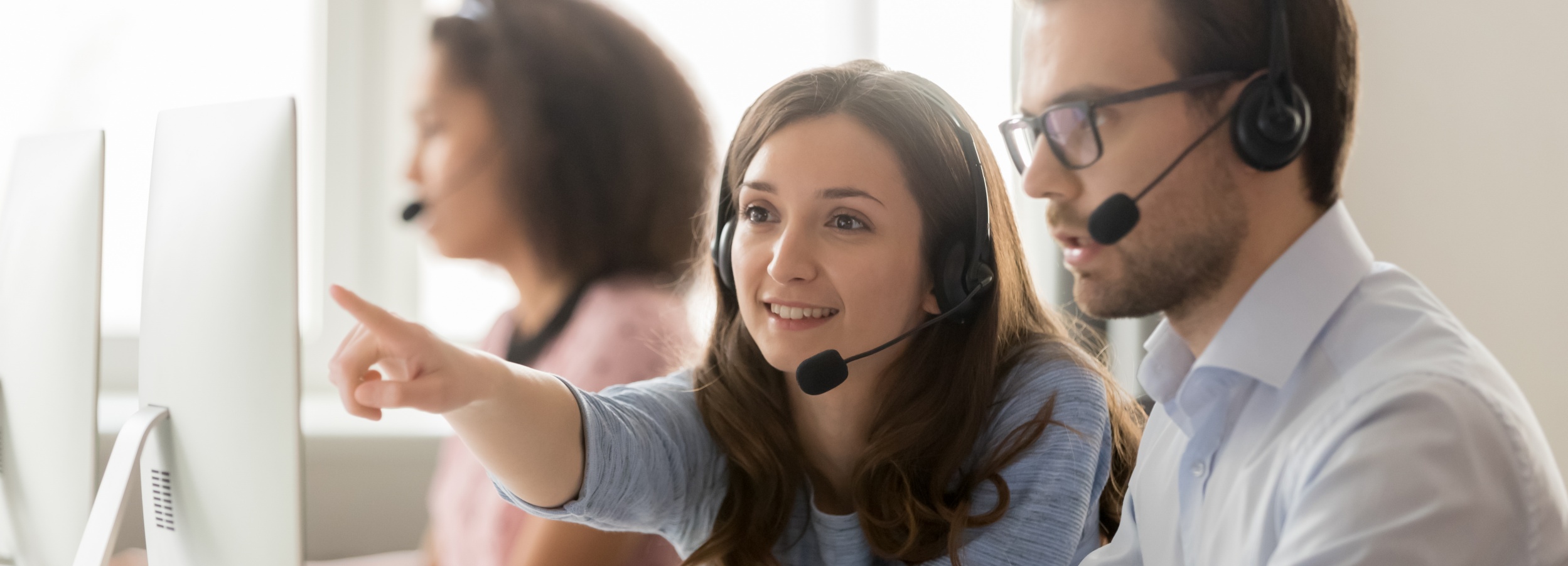 Das Bild zeigt eine Gruppe von Kundendienstmitarbeitern bei der Arbeit, wobei sich ein Mann und eine Frau in den Vordergrund rücken, während sie auf einen Computerbildschirm schauen und Anweisungen geben.
Das Bild zeigt drei Kundendienstmitarbeiter, die alle Headsets tragen und vor Computern sitzen. Im Vordergrund sind eine Frau und ein Mann zu sehen. Die Frau zeigt mit dem Finger auf den Computerbildschirm und lächelt, während der Mann aufmerksam zuhört. Beide tragen formelle Kleidung: Die Frau ein hellblaues Hemd und der Mann ein weißes Hemd mit Brille. Im Hintergrund ist eine weitere Mitarbeiterin zu sehen, ebenfalls mit Headset, aber unscharf. Die Atmosphäre wirkt professionell und kooperativ, mit einem positiven und hilfsbereiten Unterton. Die Beleuchtung ist hell und gleichmäßig, was die Gesichter der Mitarbeiter gut sichtbar macht. Die Farbtöne sind sanft und beruhigend, was eine angenehme Arbeitsumgebung suggeriert.