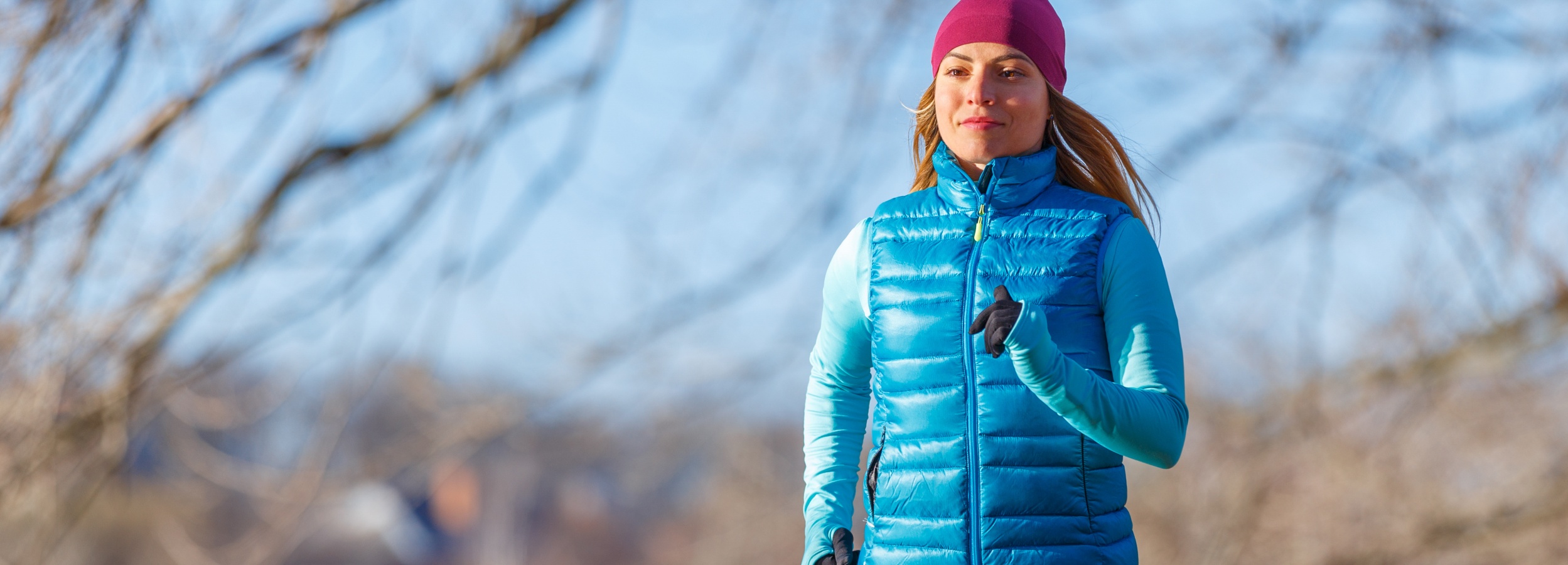 Das Bild zeigt eine Frau mittleren Alters, die im Freien joggt und dabei sportliche Kleidung trägt.
Im Detail zeigt das Bild eine Frau, die sich beim Joggen oder Laufen im Freien befindet. Sie trägt eine purpurrote Mütze, eine türkisfarbene, gesteppte Weste und ein langärmeliges Oberteil in einem ähnlichen Farbton. Ihre Hände sind mit schwarzen Handschuhen bedeckt. Die Frau scheint sich in einem Park oder einer ähnlichen Grünanlage zu befinden, da im Hintergrund Bäume und ein blauer Himmel erkennbar sind. Die Äste der Bäume sind kahl, was auf eine kühlere Jahreszeit hindeutet. Das Licht ist hell und sonnig, was eine positive und dynamische Atmosphäre erzeugt. Die Frau wirkt fokussiert und entschlossen, was die Stimmung von Aktivität und Gesundheit unterstreicht.
