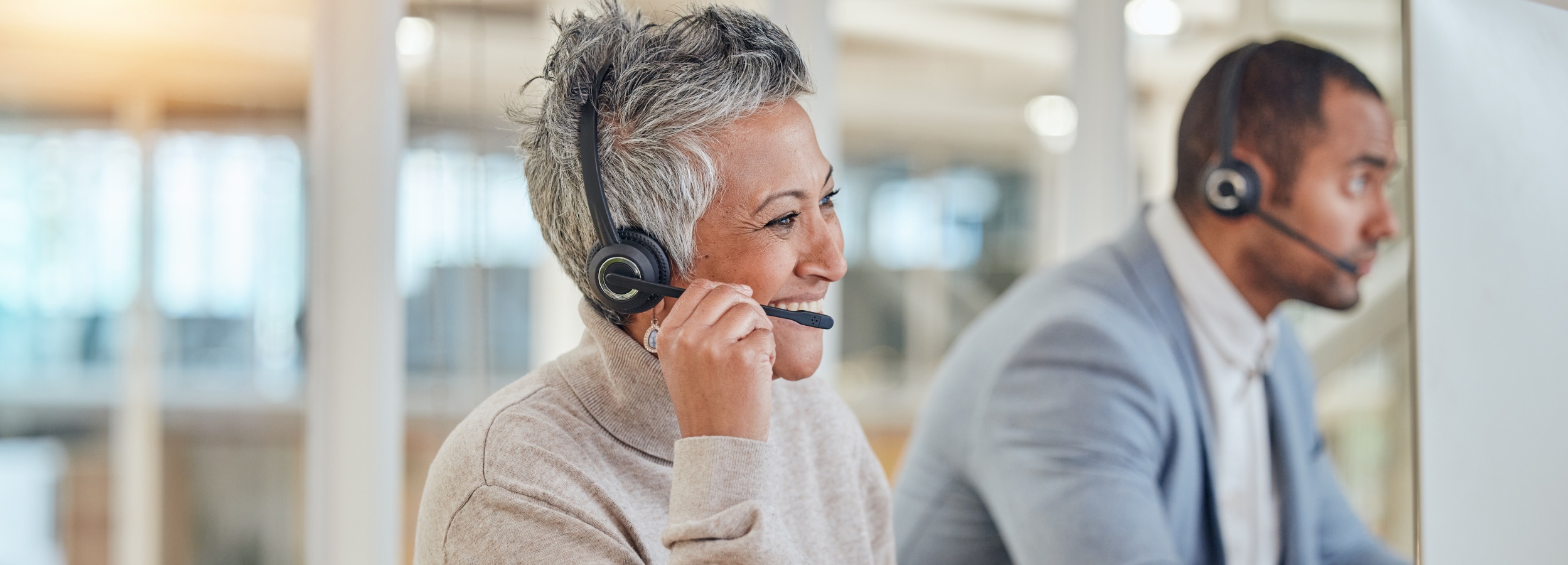 Das Bild zeigt zwei Callcenter-Mitarbeiter, die lächelnd und konzentriert miteinander interagieren und dabei Headsets tragen.

Im Vordergrund des Bildes ist eine Frau mittleren Alters mit kurzen, grauen Haaren zu sehen, die leicht zum Betrachter gedreht ist und in ihr Headset spricht. Sie trägt ein beiges Rollkragenoberteil und eine auffällige, ornamentale Ohrring. Ihr Gesichtsausdruck ist von einem freundlichen Lächeln und einer offensichtlichen Freude an ihrer Tätigkeit geprägt, ihre Augen sind leicht zusammengekniffen, was auf eine positive Interaktion hindeutet. Ihre linke Hand nähert sich dem Mikrofon ihres Headsets, was darauf hindeutet, dass sie gerade spricht oder eine Unterbrechung erwägt.

Im Hintergrund und leicht unscharf befindet sich ein Mann, der ebenfalls ein Headset trägt und seinem Gegenüber aufmerksam zuzuhören scheint. Er ist mit einem hellblauen Sakko über einem weißen Hemd bekleidet und blickt nach rechts, möglicherweise auf einen Bildschirm oder zu seinem Gegenüber. Sein Gesichtsausdruck ist konzentriert und professionell.

Die Atmosphäre des Bildes ist hell und professionell, mit weichem Licht, das von links einfällt und eine warme Ausleuchtung schafft. Der Hintergrund ist unscharf, aber es scheint sich um ein modernes Büroumfeld mit Fenstern oder Glasabtrennungen zu handeln. Die Stimmung ist positiv und produktiv, was die Zufriedenheit und das Engagement der dargestellten Personen vermittelt. Es suggeriert ein positives Arbeitsumfeld, in dem Kommunikation und Kundenservice im Vordergrund stehen.
