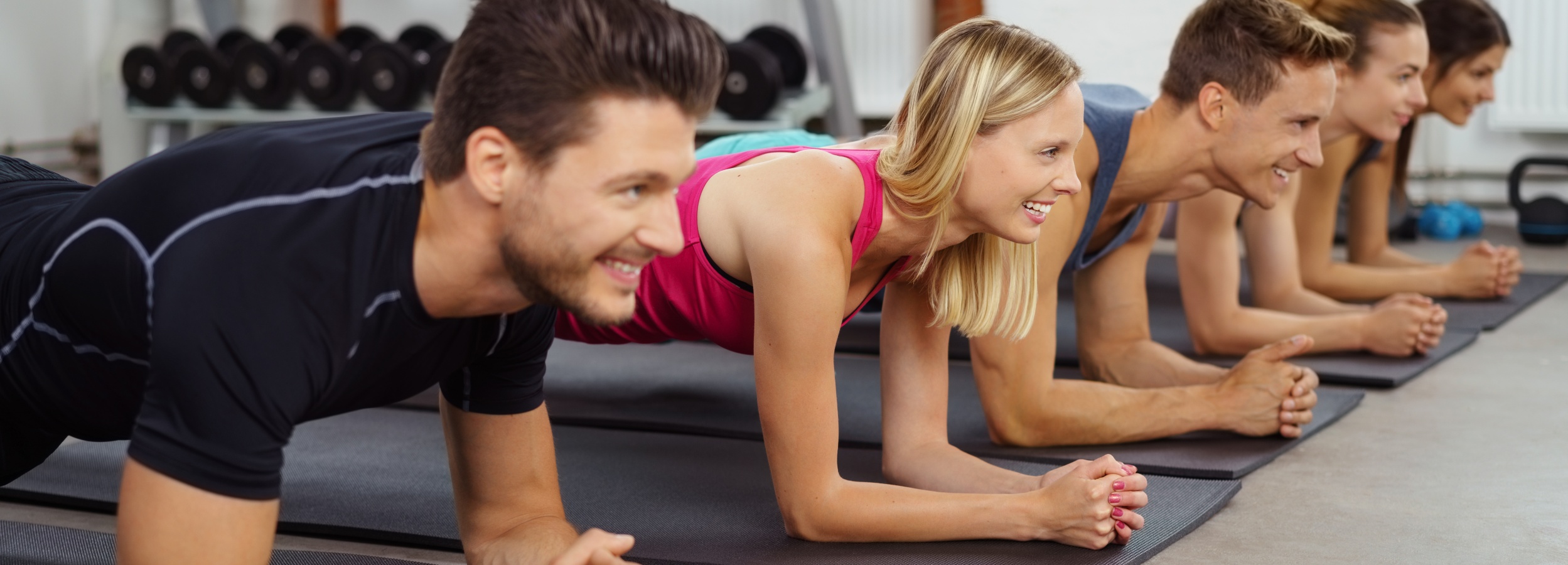 Gruppe junger Leute in Sportsachen trainiert im Studio auf Gymnastikmatten und stützt sich dabei auf den Unterarmen ab.