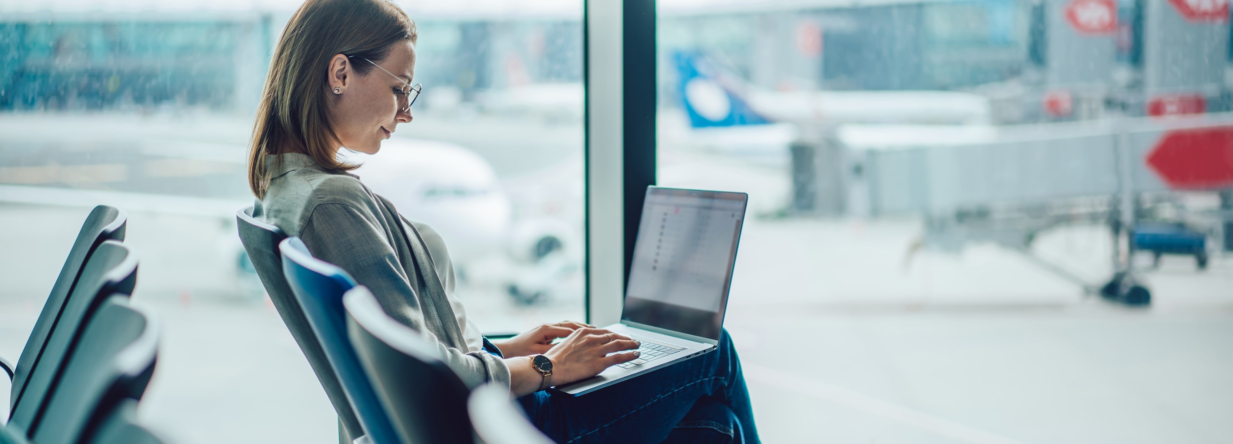 Das Bild zeigt eine Frau, die konzentriert an ihrem Laptop in einem Flughafen-Wartezimmer arbeitet.
Im Bild ist eine junge Frau mit braunen, schulterlangen Haaren zu sehen, die eine graue Bluse und Jeans trägt. Sie trägt eine Brille und eine Uhr am Handgelenk. Sie sitzt auf einem bequemen Stuhl in einem Flughafen-Wartezimmer und arbeitet an ihrem Laptop, der auf ihrem Schoß liegt. Ihre Hände tippen konzentriert auf die Tastatur. Ihr Blick ist auf den Bildschirm gerichtet und ihr Gesichtsausdruck wirkt fokussiert und konzentriert. Im Hintergrund ist durch ein großes Fenster die Rollbahn eines Flughafens mit Flugzeugen und Gepäckwagen unscharf zu sehen. Die Atmosphäre ist ruhig und konzentriert, obwohl der Ort ein geschäftiger Flughafen ist. Die Farben sind gedämpft, die Lichtverhältnisse hell und natürlich. Der gesamte Eindruck ist einer von konzentrierter Arbeit in einer öffentlichen, aber ruhigen Umgebung.