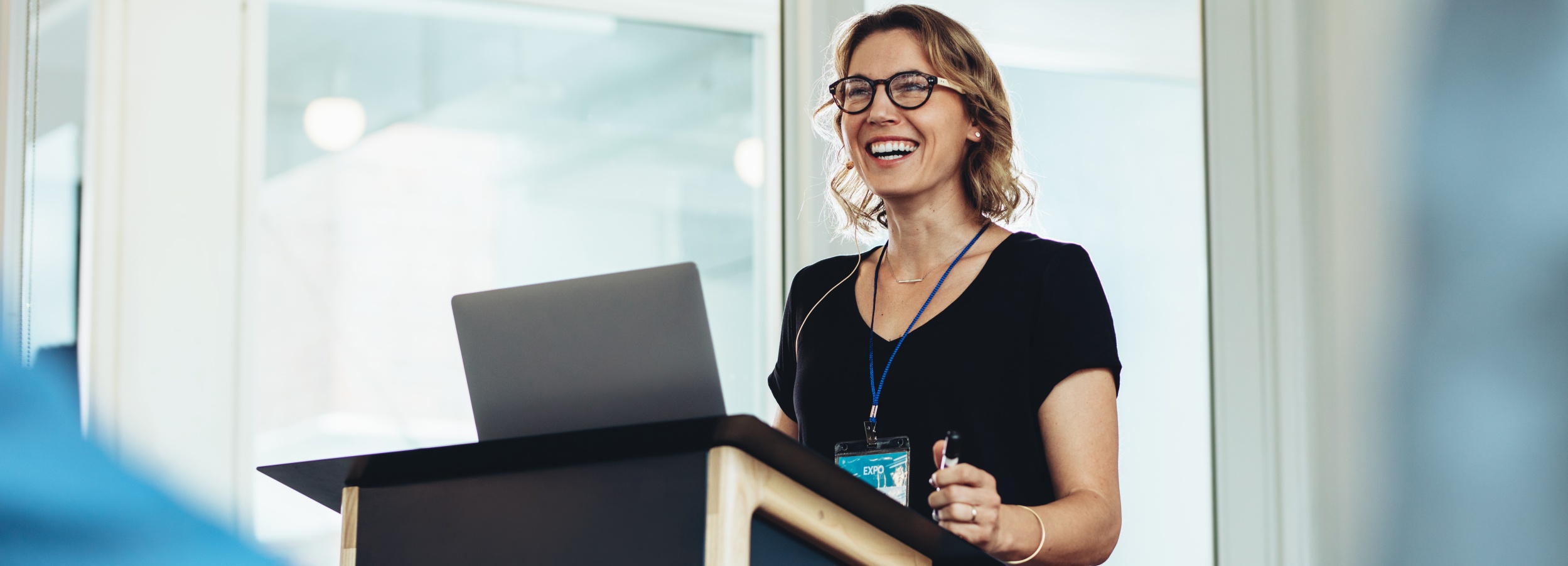Eine lachende Frau steht hinter einem Podium, auf dem ein Laptop steht.
Das Bild zeigt eine Frau mittleren Alters mit blonden, schulterlangen Haaren und einer Brille mit dicker, dunkler Fassung. Sie trägt ein schwarzes Oberteil mit V-Ausschnitt und eine Kette mit einem kleinen, horizontalen Anhänger. Um ihren Hals hängt ein Lanyard mit einem Namensschild, auf dem "EXPO" steht. Sie steht hinter einem dunklen Pult, das eine schräge, helle Holzfront hat. Auf dem Pult steht ein grauer Laptop, dessen Bildschirm leicht geöffnet ist. Die Frau hält einen schwarzen Stift in ihrer rechten Hand und lacht herzlich, wobei ihre Augen geschlossen sind und sie den Kopf leicht nach hinten neigt. Ihre linke Hand ruht auf dem Pult. Im Hintergrund ist eine helle, diffuse Umgebung zu sehen, die durch große Fenster oder Glasflächen erhellt wird. Es herrscht eine positive, lebhafte und professionelle Atmosphäre, die durch das Lachen und die offene Körpersprache der Frau vermittelt wird.