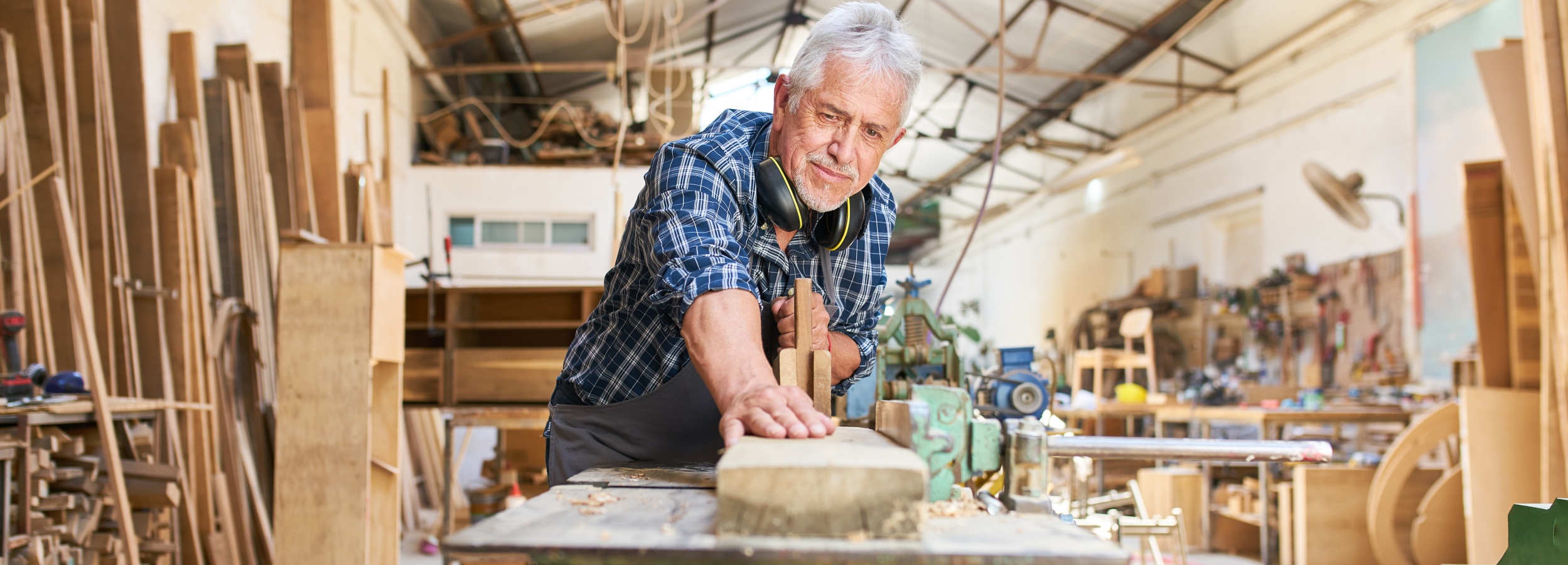 Ein älterer Handwerker arbeitet konzentriert in seiner Werkstatt an einem feinen Holzstück.
Das Bild zeigt einen älteren Mann mit grauem Haar und einem karierten Hemd, der in einer Werkstatt steht und ein Holzstück bearbeitet. Er trägt Kopfhörer um den Hals. Seine Hände sind auf dem Holzstück und einem Stechbeitel platziert. Im Hintergrund sind Regale voller Holzstücke und Werkzeuge zu sehen. Die Werkstatt wirkt belebt und voller Arbeitsmaterialien. Die Atmosphäre ist handwerklich und professionell, geprägt von der Hingabe des Mannes zu seinem Handwerk.