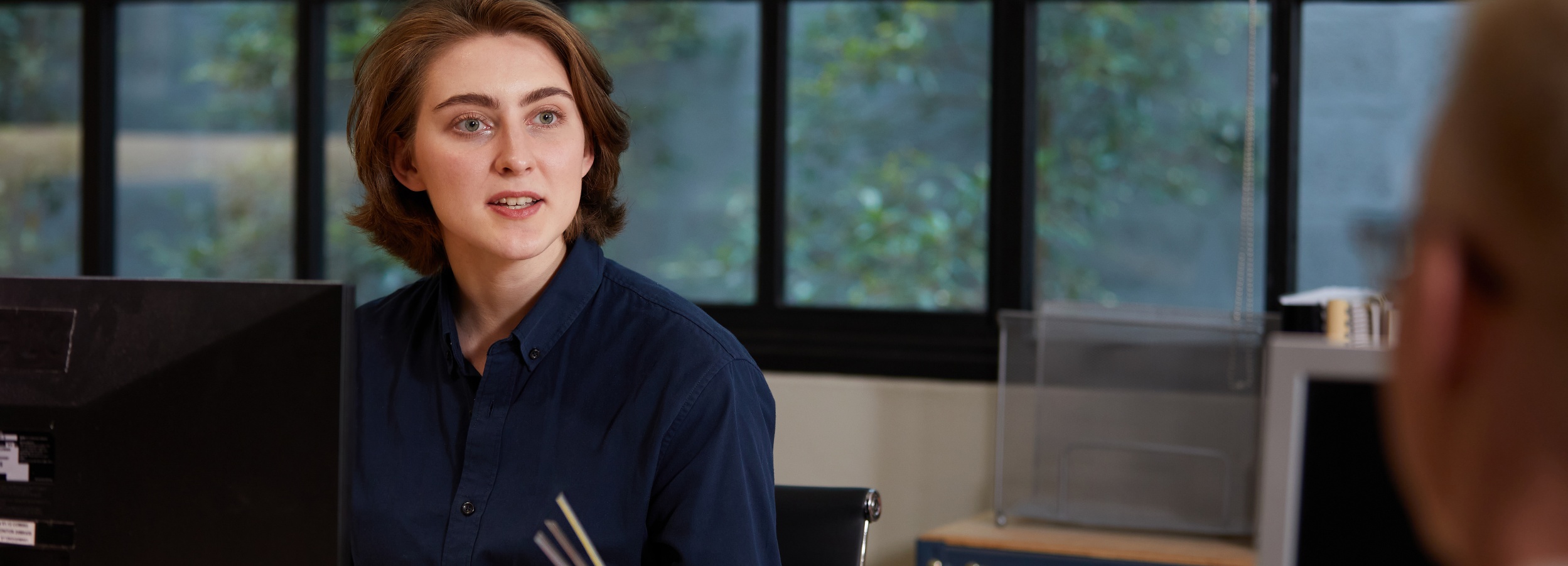 Das Bild zeigt eine junge Frau in einem Büro, die mit einem Kollegen spricht.

Im Detail zeigt das Bild den Oberkörper einer jungen Frau mit hellbraunen, schulterlangen Haaren, die ein dunkelblaues Hemd trägt. Sie sitzt an einem Schreibtisch, vor dem ein schwarzer Computermonitor steht. Ihr Blick ist nach rechts gewandt, wo man nur den Hinterkopf einer anderen blondhaarigen Person sieht, was darauf hindeutet, dass sie sich in einem Gespräch befindet. Im Hintergrund ist ein Fenster zu sehen, das einen Blick ins Grüne freigibt, was dem Raum eine beruhigende Atmosphäre verleiht. Auf der rechten Seite des Bildes sind Teile eines weiteren Schreibtisches mit Bürobedarf zu erkennen. Die Atmosphäre wirkt professionell und gleichzeitig entspannt, was durch das natürliche Licht und die Pflanzen im Hintergrund verstärkt wird.