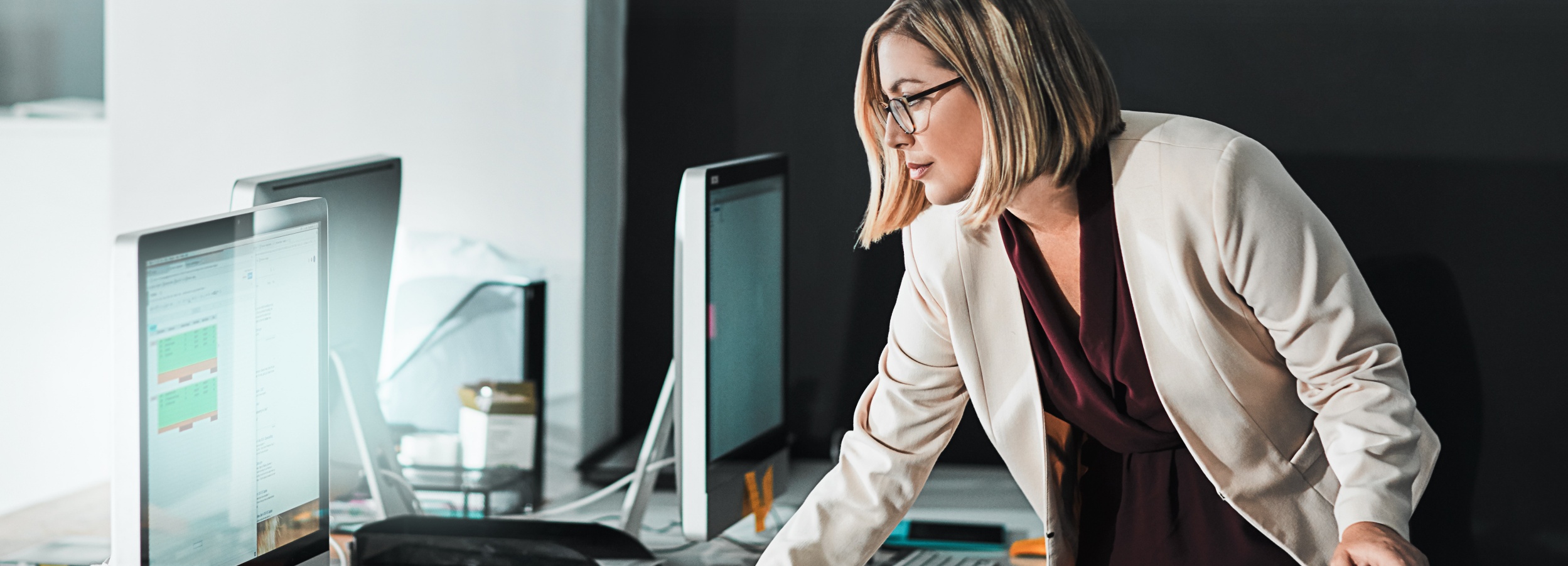 Eine professionell gekleidete Frau beugt sich über einen Schreibtisch voller Computerbildschirme und anderer Bürogegenstände, während sie konzentriert auf einen der Bildschirme schaut.
Das Bild zeigt eine blonde Frau mit Brille, die einen beigen Blazer und eine dunkelrote Bluse trägt. Sie steht an einem Schreibtisch, auf dem mehrere Computerbildschirme stehen. Auf den Bildschirmen sind verschiedene Fenster und Anwendungen zu sehen. Die Frau scheint sich auf den Inhalt eines der Bildschirme zu konzentrieren. Die Atmosphäre des Bildes ist professionell und konzentriert. Der Hintergrund ist unscharf und dunkel, was den Fokus auf die Frau und die Computerbildschirme lenkt. Das Bild vermittelt den Eindruck von harter Arbeit und Engagement im Büro.