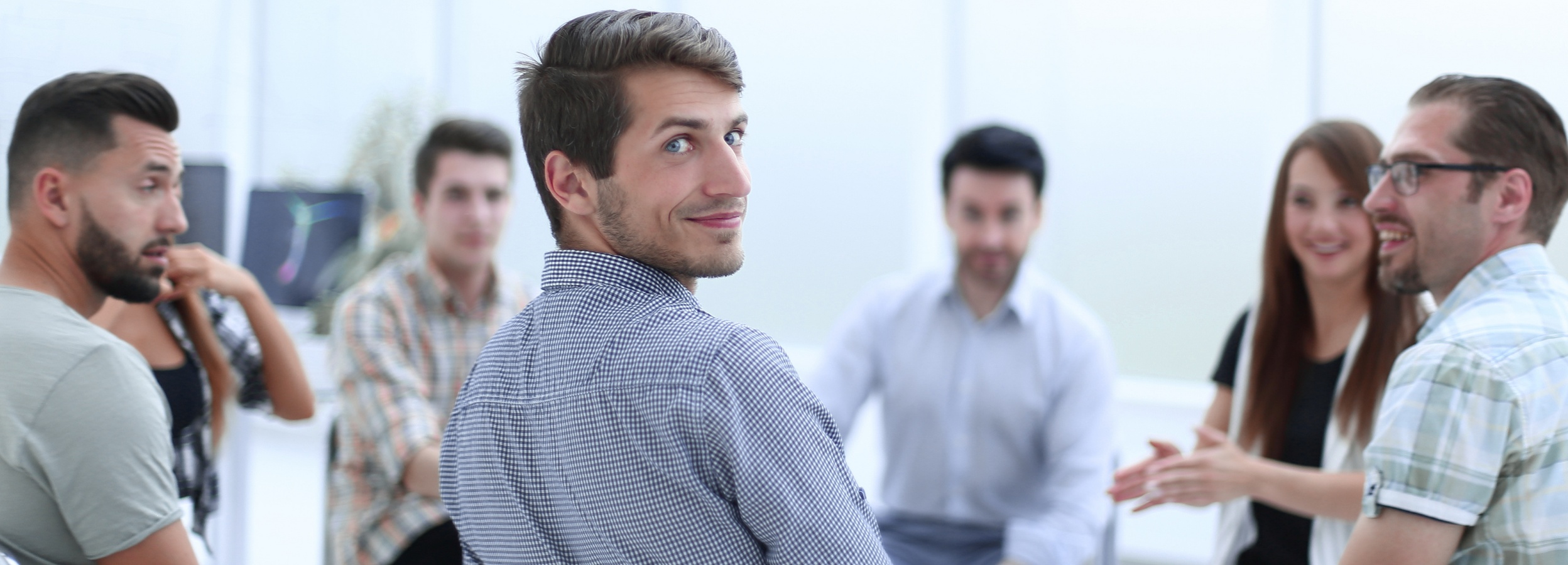 Das Bild zeigt eine Gruppe von Menschen in einem Büro, wobei ein Mann im Vordergrund hervorsticht, der über seine Schulter lächelt.

Im Detail zeigt das Bild eine Gruppe von etwa sieben Personen, die sich in einem hellen, modernen Büroumfeld befinden. Der Fokus liegt auf einem Mann im Vordergrund, der ein blau-weiß kariertes Hemd trägt und direkt in die Kamera lächelt, während er über seine Schulter schaut. Er hat dunkles Haar und blaue Augen, und sein Gesichtsausdruck wirkt freundlich und einladend.

Um ihn herum befindet sich der Rest der Gruppe, die sich scheinbar in einer Diskussion oder einem Meeting befindet. Die Personen sind teilweise unscharf, was darauf hindeutet, dass der Fokus auf dem Mann im Vordergrund liegt. Sie tragen verschiedene Arten von Kleidung, darunter Hemden, Blusen und T-Shirts. Einige von ihnen lächeln oder scheinen aktiv an der Unterhaltung teilzunehmen. Im Hintergrund sind Büromöbel und Computer zu erkennen, was den Eindruck eines geschäftigen Arbeitsplatzes verstärkt.

Die Atmosphäre des Bildes wirkt positiv und dynamisch, was durch die lächelnden Gesichter und die offene Körpersprache der Personen unterstrichen wird. Die Stimmung ist entspannt und kooperativ, was auf ein gutes Arbeitsklima hindeutet.