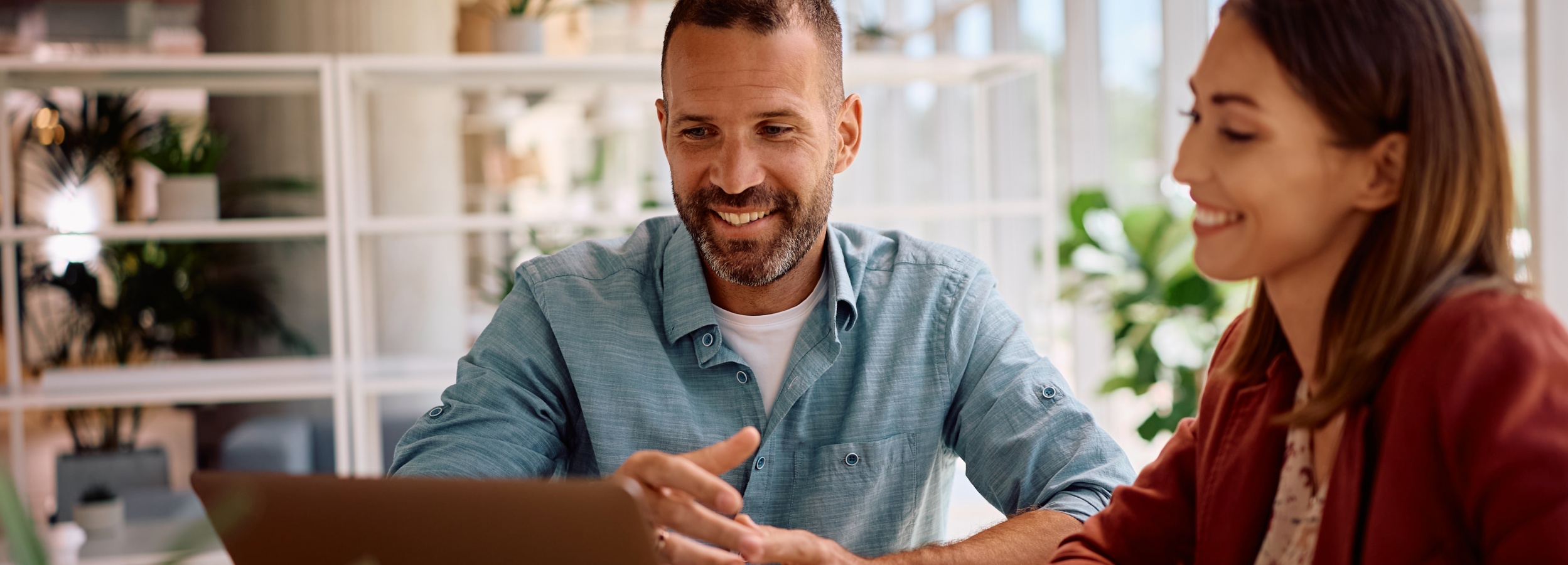 Das Bild zeigt einen Mann und eine Frau, die an einem Tisch vor einem Laptop sitzen und sich angeregt unterhalten.

Im Vordergrund sind ein Mann und eine Frau zu sehen, die an einem Tisch sitzen. Der Mann, zu seiner Rechten, hat einen leichten Bart und trägt ein hellblaues Hemd über einem weißen T-Shirt. Er lächelt und gestikuliert mit seinen Händen, was darauf hindeutet, dass er etwas erklärt oder betont. Die Frau, zu seiner Linken, hat braune Haare und trägt eine rostrote Jacke über einem gemusterten Oberteil. Sie lächelt ebenfalls und scheint aufmerksam zuzuhören.

Zwischen den beiden Personen befindet sich ein Laptop, der auf dem Tisch steht. Vor dem Laptop liegt ein Blatt Papier. Der Tisch selbst scheint aus Holz zu sein.

Im Hintergrund ist ein Raum zu sehen, der hell und modern wirkt. Ein Regal mit Pflanzen und dekorativen Elementen ist erkennbar. Durch das Fenster im Hintergrund sind verschwommene Umrisse von Gebäuden und Fahrzeugen zu erkennen, was darauf hindeutet, dass sich die Szene in einem urbanen Umfeld abspielt.

Die Atmosphäre des Bildes ist positiv und professionell. Die Stimmung ist freundlich und aufmerksam. Die Interaktion zwischen den beiden Personen deutet auf ein produktives Gespräch oder eine Zusammenarbeit hin. Das helle und moderne Umfeld verstärkt den Eindruck einer positiven und dynamischen Arbeitsatmosphäre.